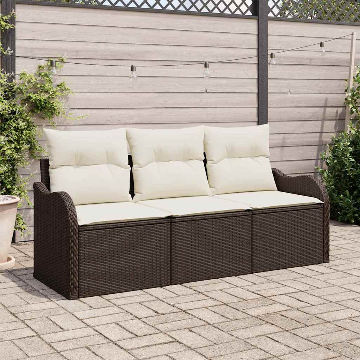 Braunes Outdoor-Sofa aus Korbgeflecht mit cremefarbenen Kissen. Es steht auf einer Steinpflaster-Terrasse mit Holzzaun und Lichterketten im Hintergrund.