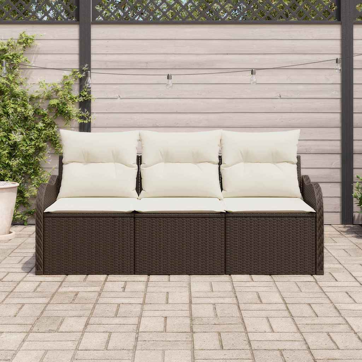 Ein braunes Korbmöbel mit cremefarbenen Kissen steht auf einer Ziegelterrasse. Im Hintergrund sind ein Holzzaun und Kletterpflanzen zu sehen.