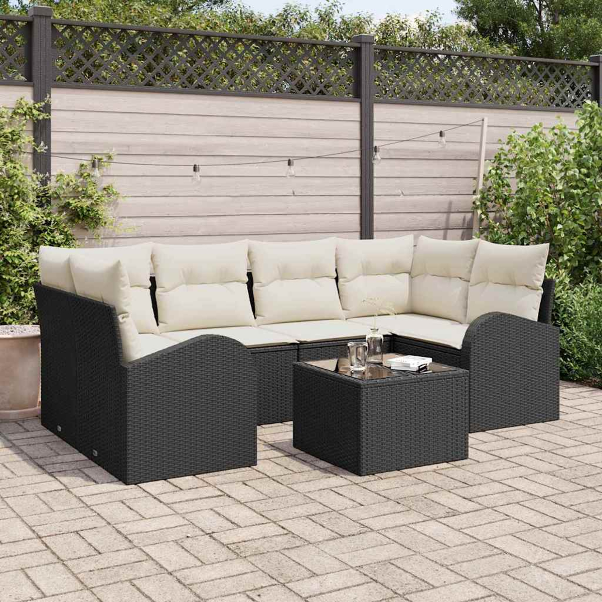 Ein schwarzes Outdoor-Sofa aus Korbgeflecht mit cremefarbenen Kissen und einem kleinen Tisch. Im Hintergrund ist ein Holzzaun mit Lichtern zu sehen.