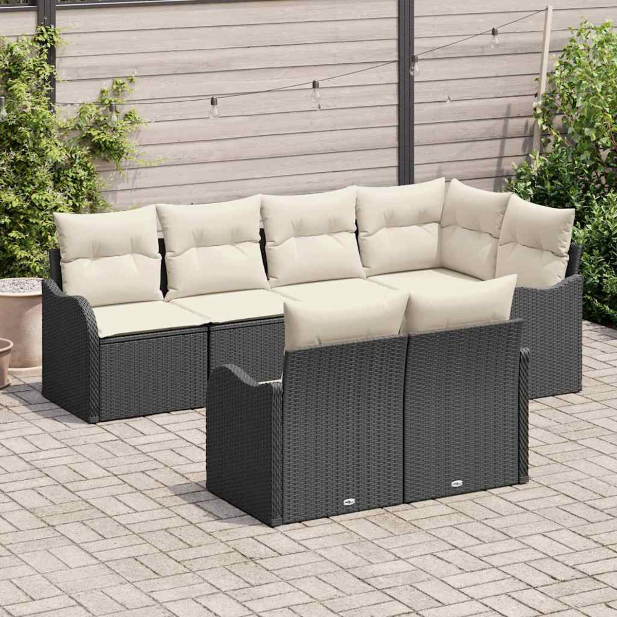 Outdoor-Ecksofa mit grauem Korbgeflecht und cremefarbenen Kissen auf einer Ziegelsteinterrasse. Holzzaun und Grün im Hintergrund.