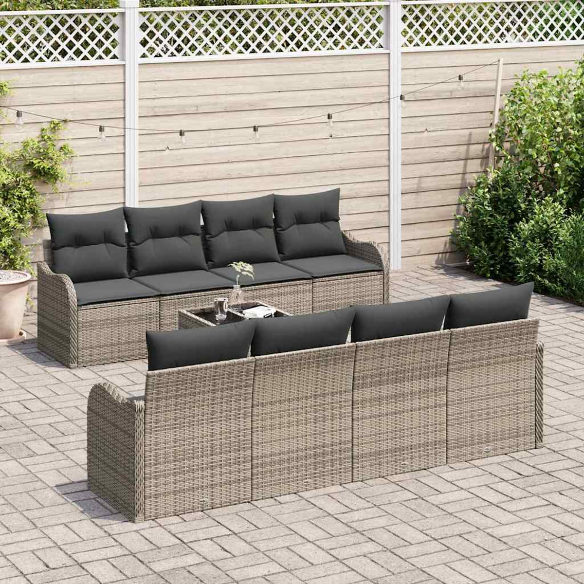 Garten-Rattan-Sofagarnitur mit grauen Kissen auf einer Backstein-Terrasse. Hintergrund mit Holzzaun, Lichterkette und grünem Laub.