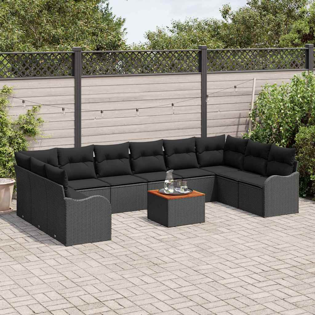 Garten-Sitzgruppe mit schwarzen Kissen, kleinem Tisch und Glaskrug auf einer Backstein-Terrasse. Holzzaun und Grün im Hintergrund.