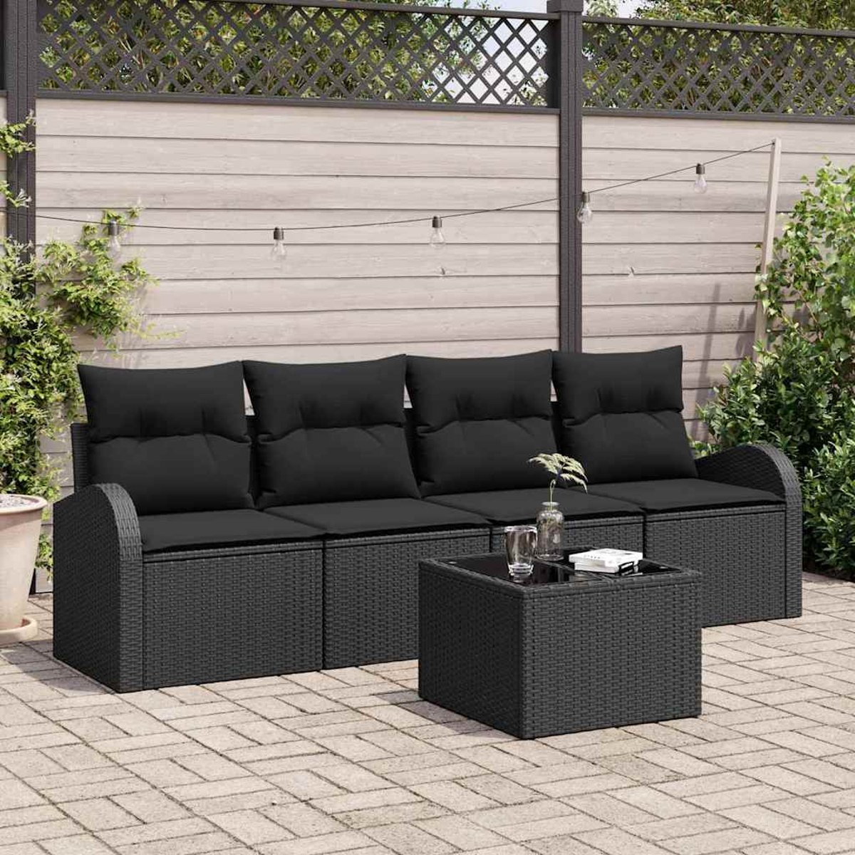 Schwarzes Outdoor-Sofa aus Korbgeflecht mit schwarzen Kissen und einem quadratischen Couchtisch auf einer gepflasterten Terrasse. Im Hintergrund ein Holzzaun.