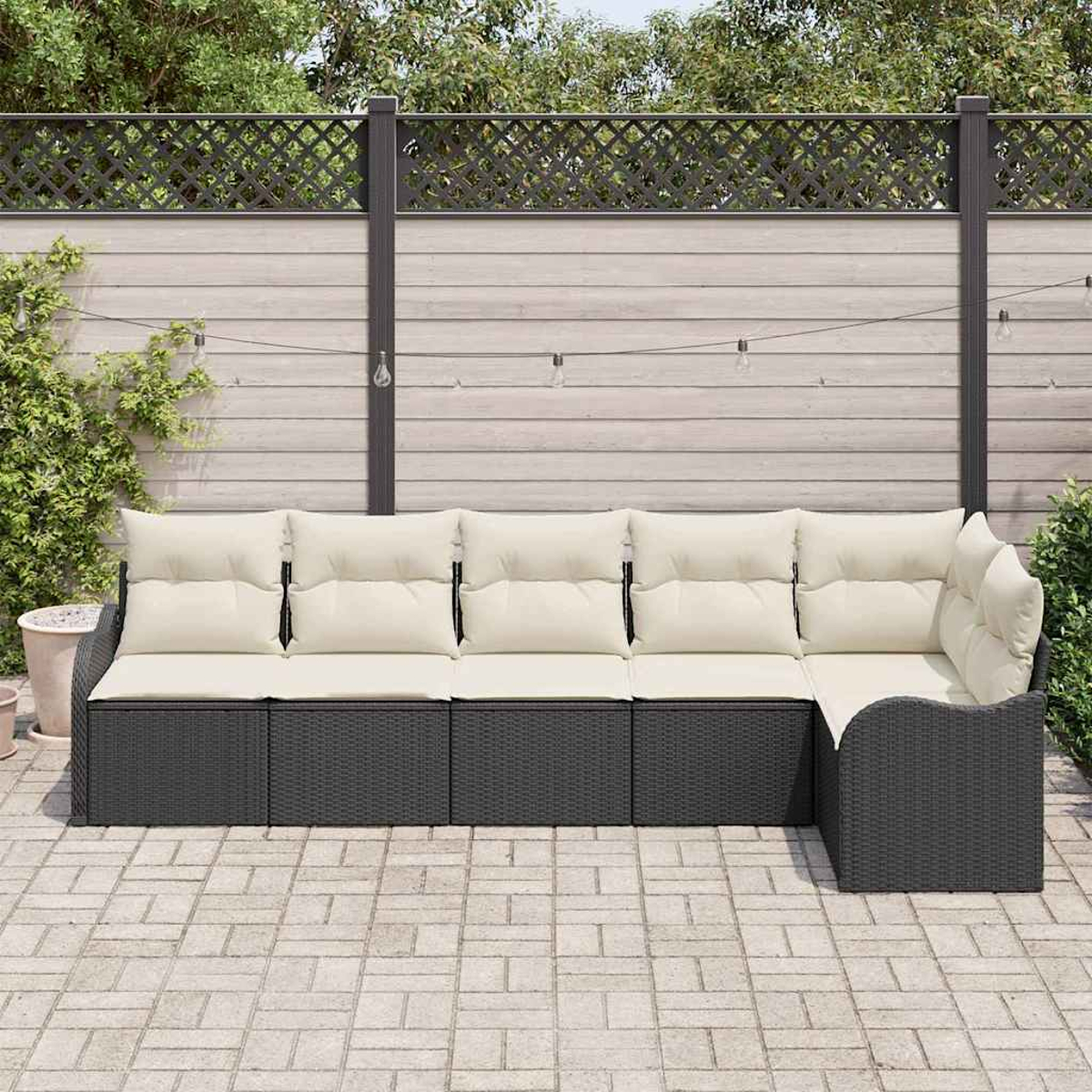 Dunkelgraues Outdoor-Sofa mit cremefarbenen Kissen steht auf einer Ziegelterrasse. Ein Holzzaun und grünes Laub im Hintergrund.