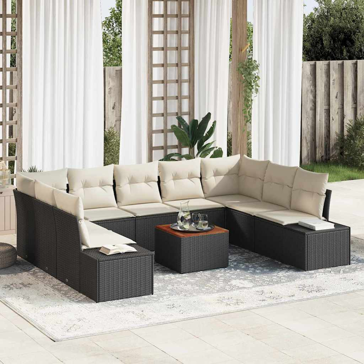 Gartensofa mit cremefarbenen Kissen und einem Couchtisch auf einem gemusterten Teppich. Weiße Vorhänge, Pflanzen und Holzzaun sind zu sehen.
