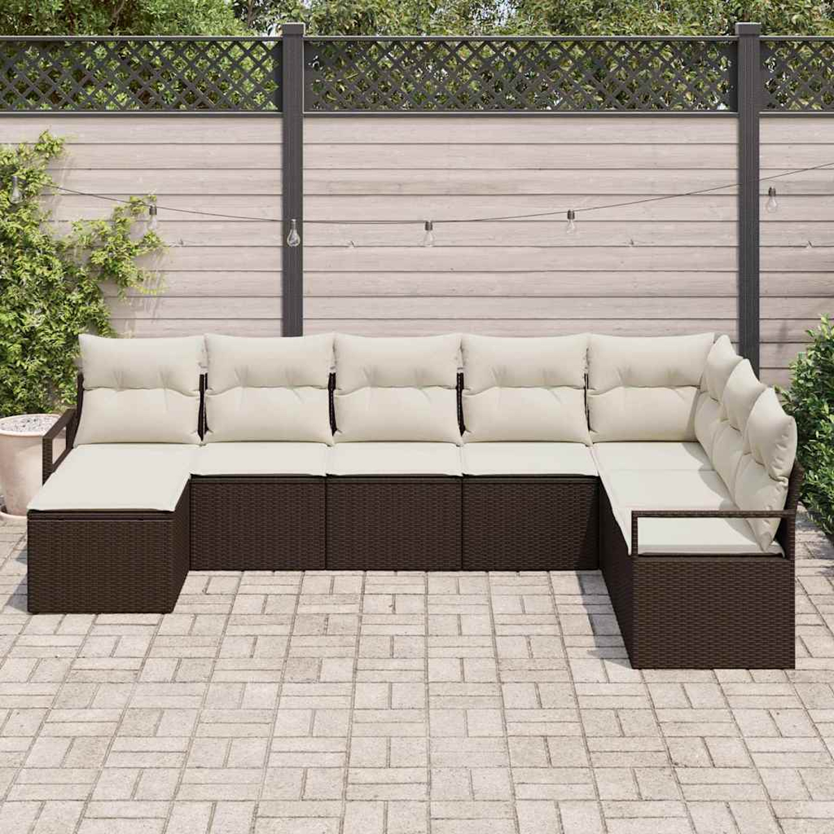 Outdoor-Ecksofa mit cremefarbenen Kissen auf braunem Korbgeflecht, auf einer Steinterrasse. Im Hintergrund ein Holzzaun und Grün.