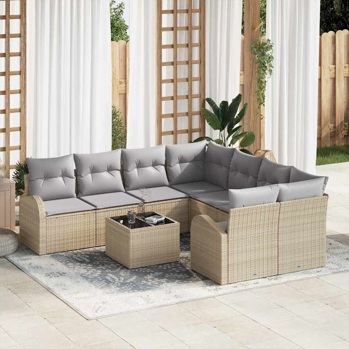 Ein Outdoor-Terrassenset mit beigen Korbmöbeln, grauen Kissen, einem Couchtisch mit Glasplatte und einem gemusterten Teppich. Weiße Vorhänge im Hintergrund.