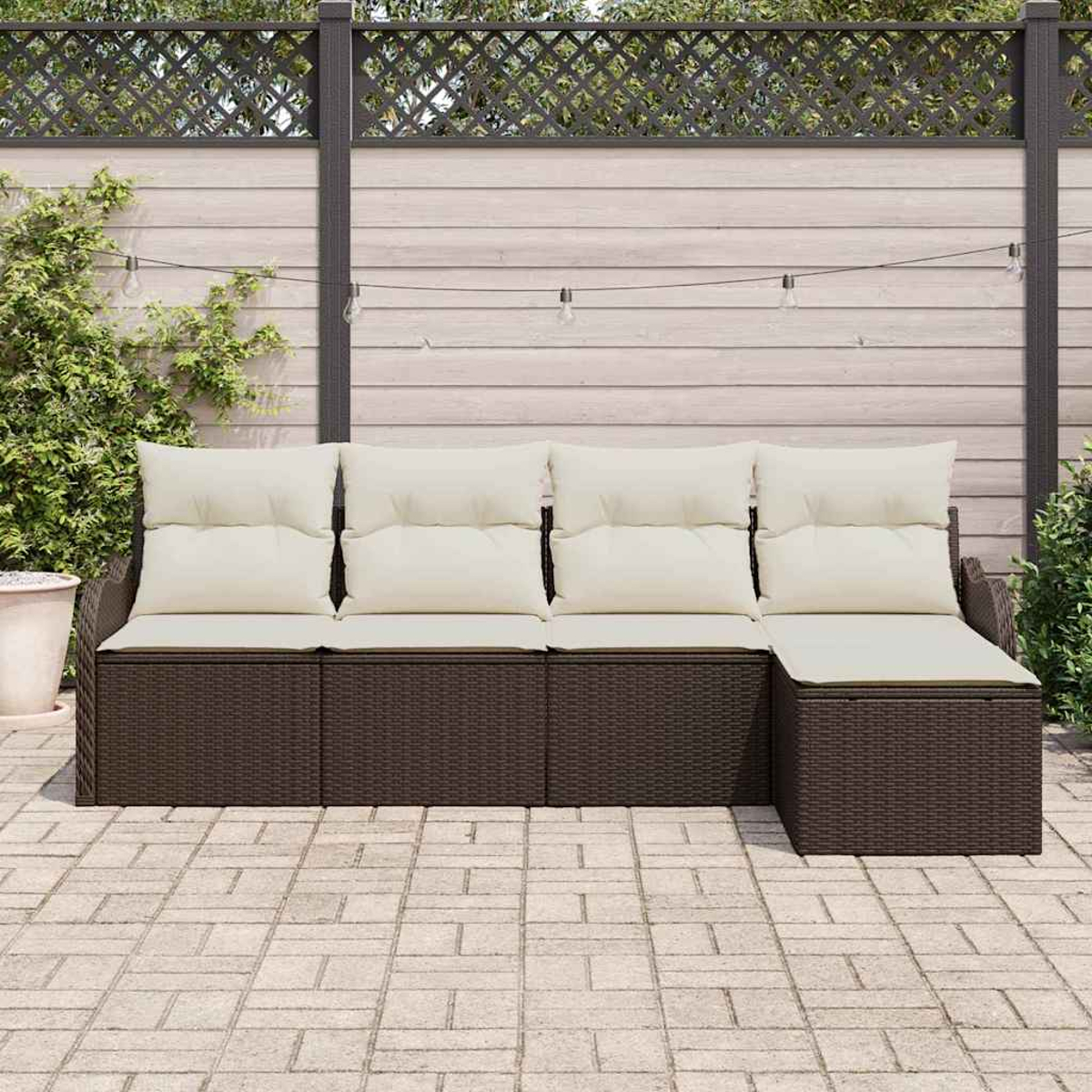Braunes Rattansofa mit cremefarbenen Kissen auf einer Ziegelterrasse. Der Hintergrund zeigt einen Holzzaun mit dekorativer Spitze und Lichterketten.