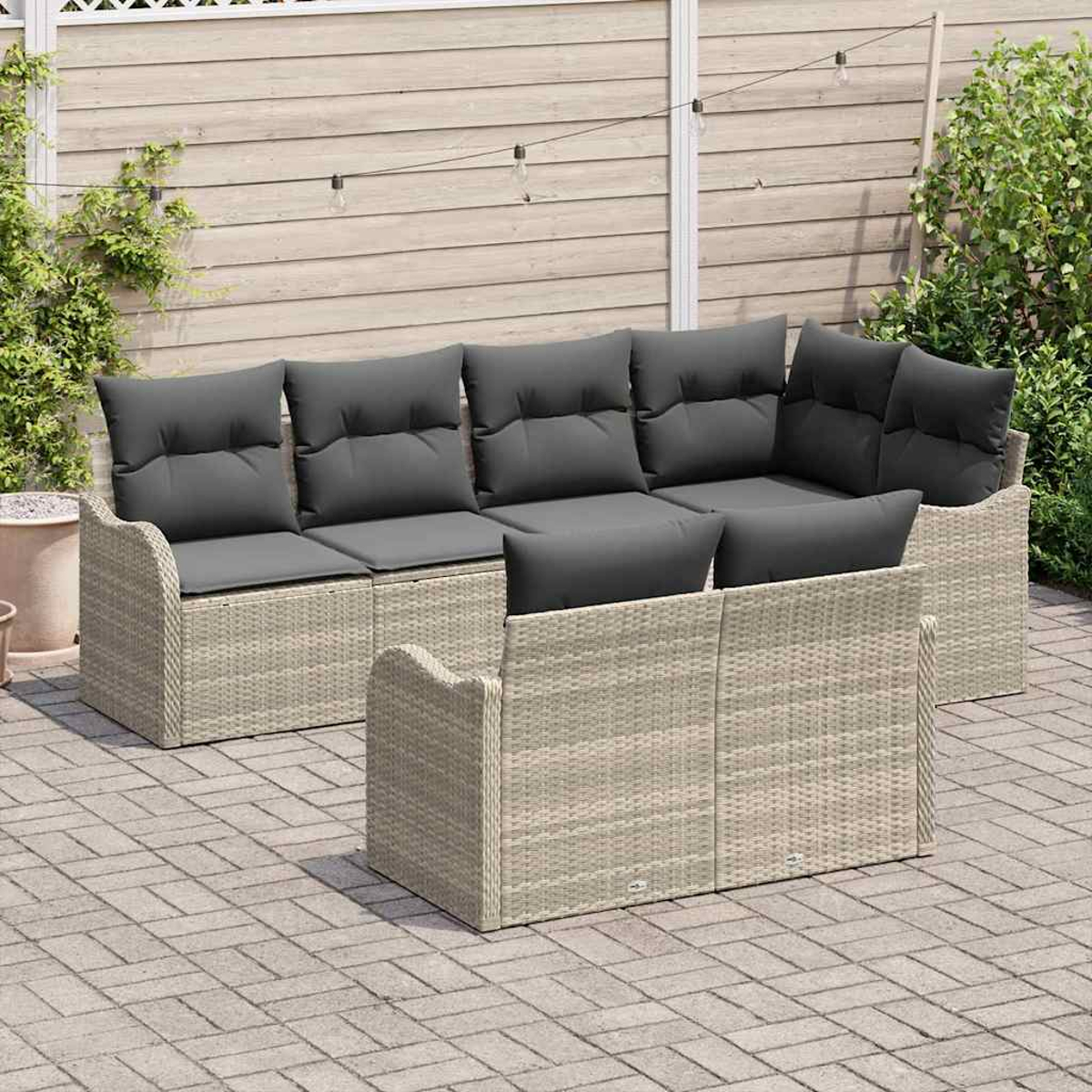 Garten-Rattansofa-Set mit grauen Kissen. Hintergrund: Holzzaun, Pflanzen und Lichterkette. Gepflasterte Terrasse im Vordergrund.