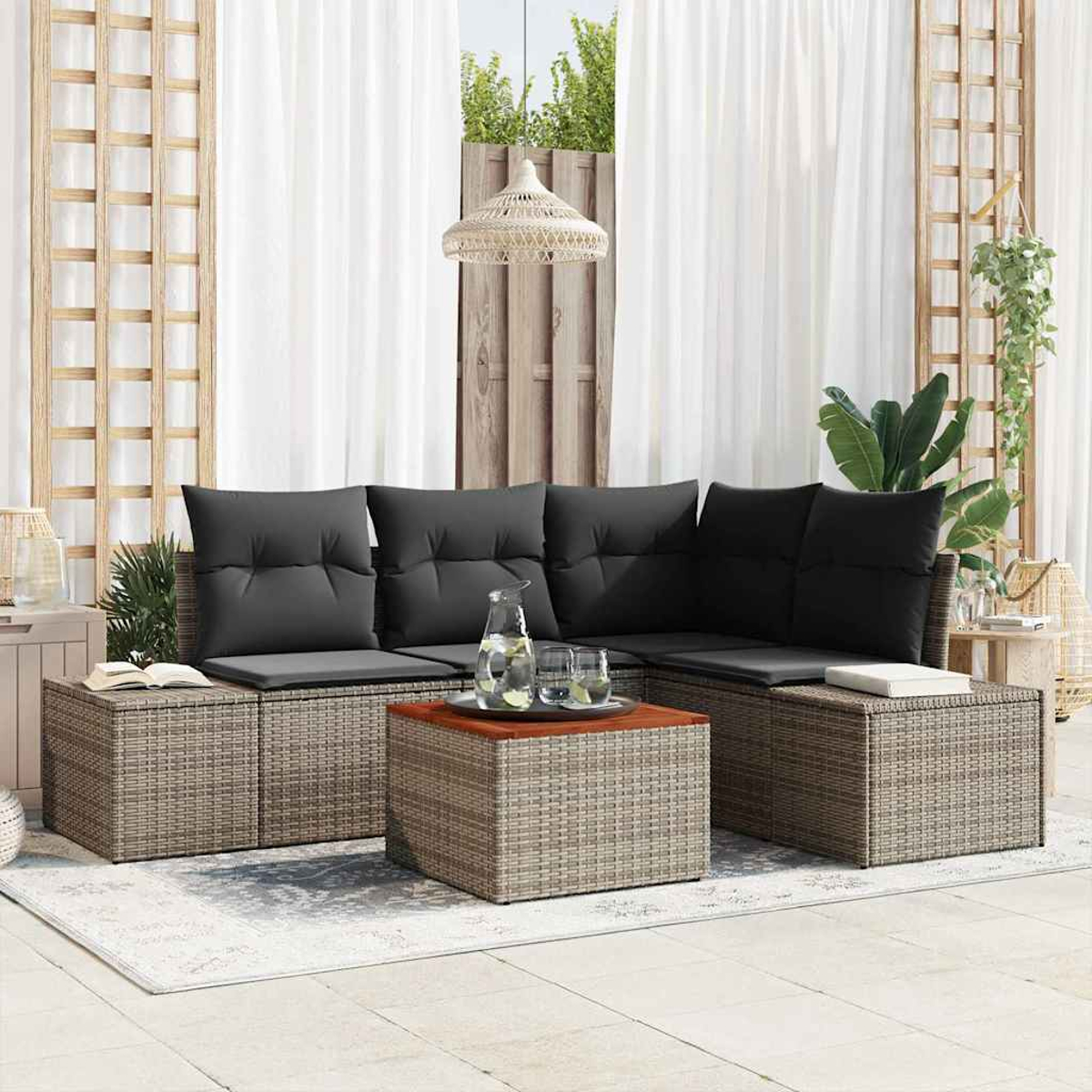 Sof á de ratán para exteriores con cojines negros, mesa de centro con agua y vasos, cortinas blancas y planta decorativa.
