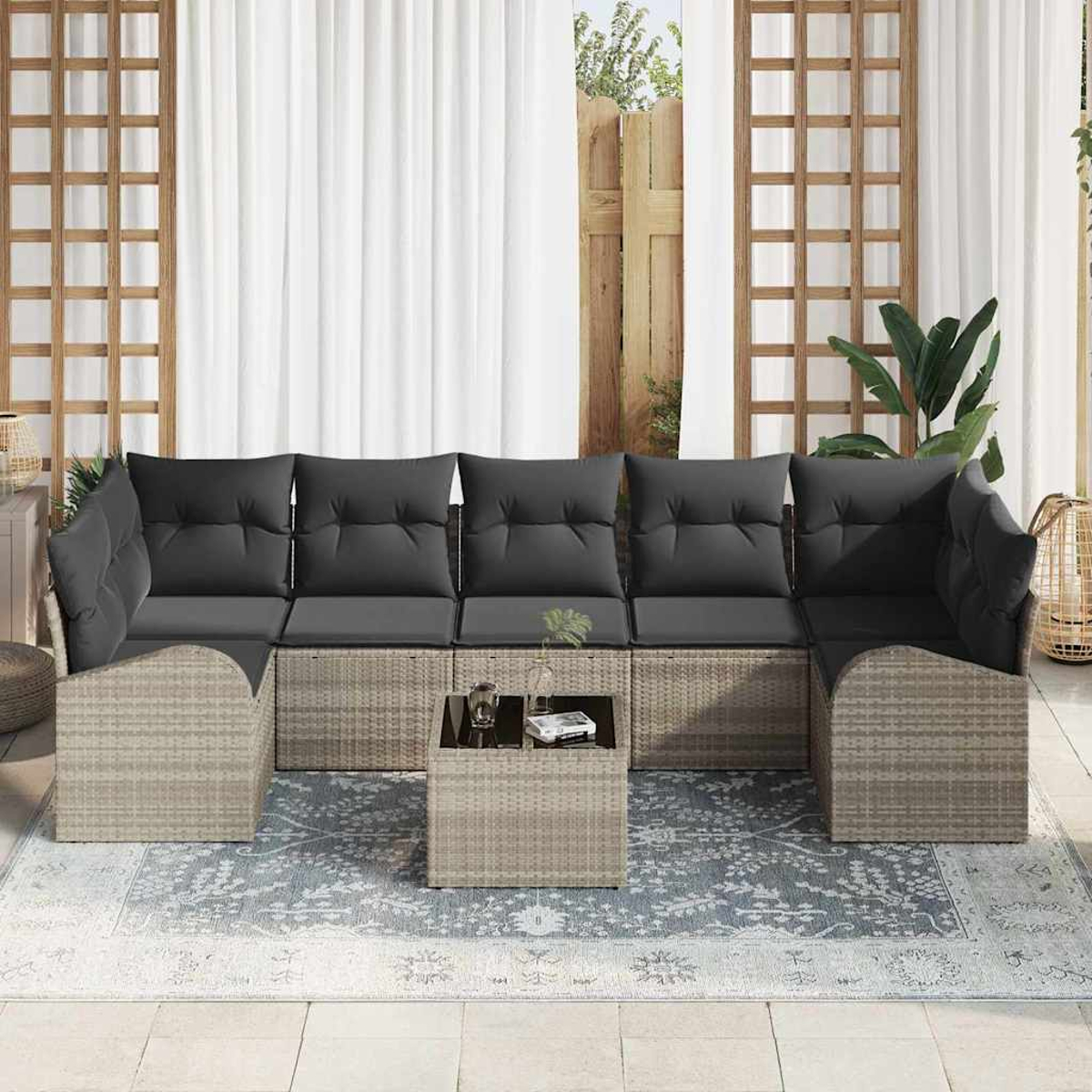 Rattansofa mit grauen Kissen auf einem gemusterten Teppich. Davor steht ein kleiner Tisch. Weiße Vorhänge und Gitter im Hintergrund.