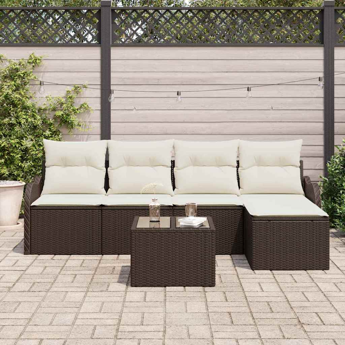Outdoor-Ecksofa mit cremefarbenen Kissen und einem kleinen Tisch. Im Hintergrund ein Zaun und Grün. Gepflasterte Terrasse im Vordergrund.