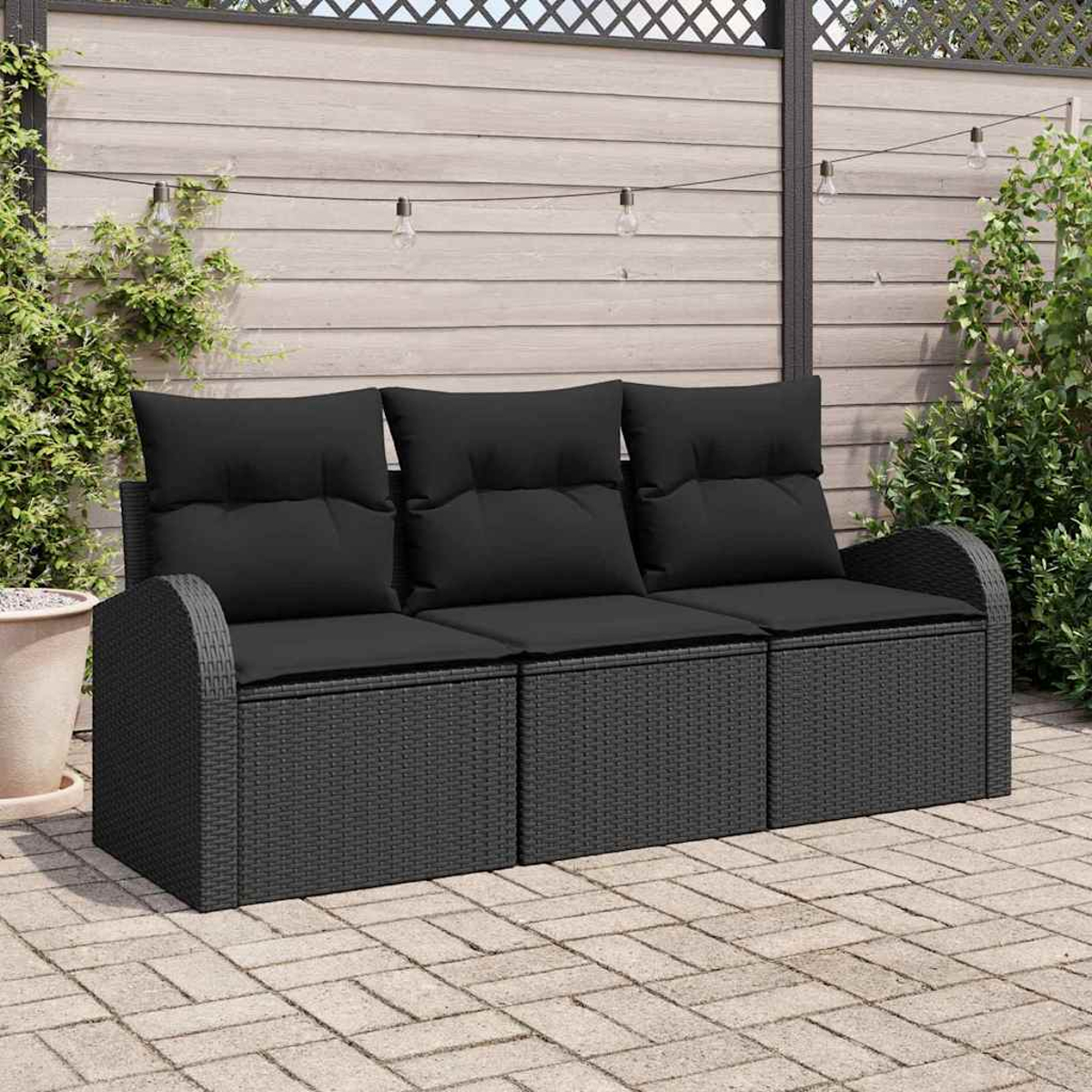 Ein schwarzes Outdoor-Sofa aus Korbgeflecht mit schwarzen Kissen. Im Hintergrund ein Holzzaun, Lichterketten und Pflanzen.