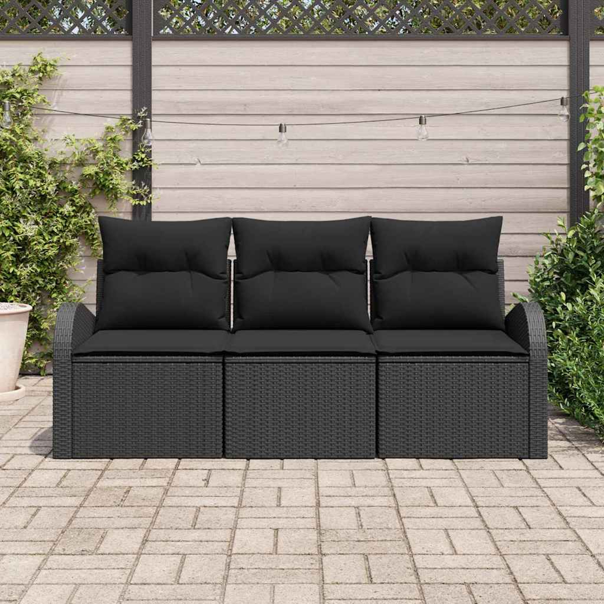 Ein schwarzes Rattan-Sofa mit Kissen steht auf einer Ziegelterrasse, im Hintergrund ein Holzzaun und Lichter.