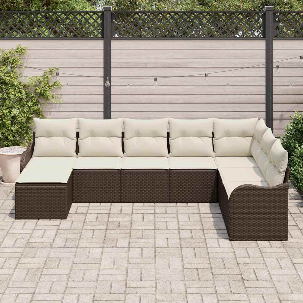 Ein braunes Korbsessel-Ecksofa mit cremefarbenen Kissen steht auf einer Steinpatio. Im Hintergrund befindet sich ein Holzzaun.