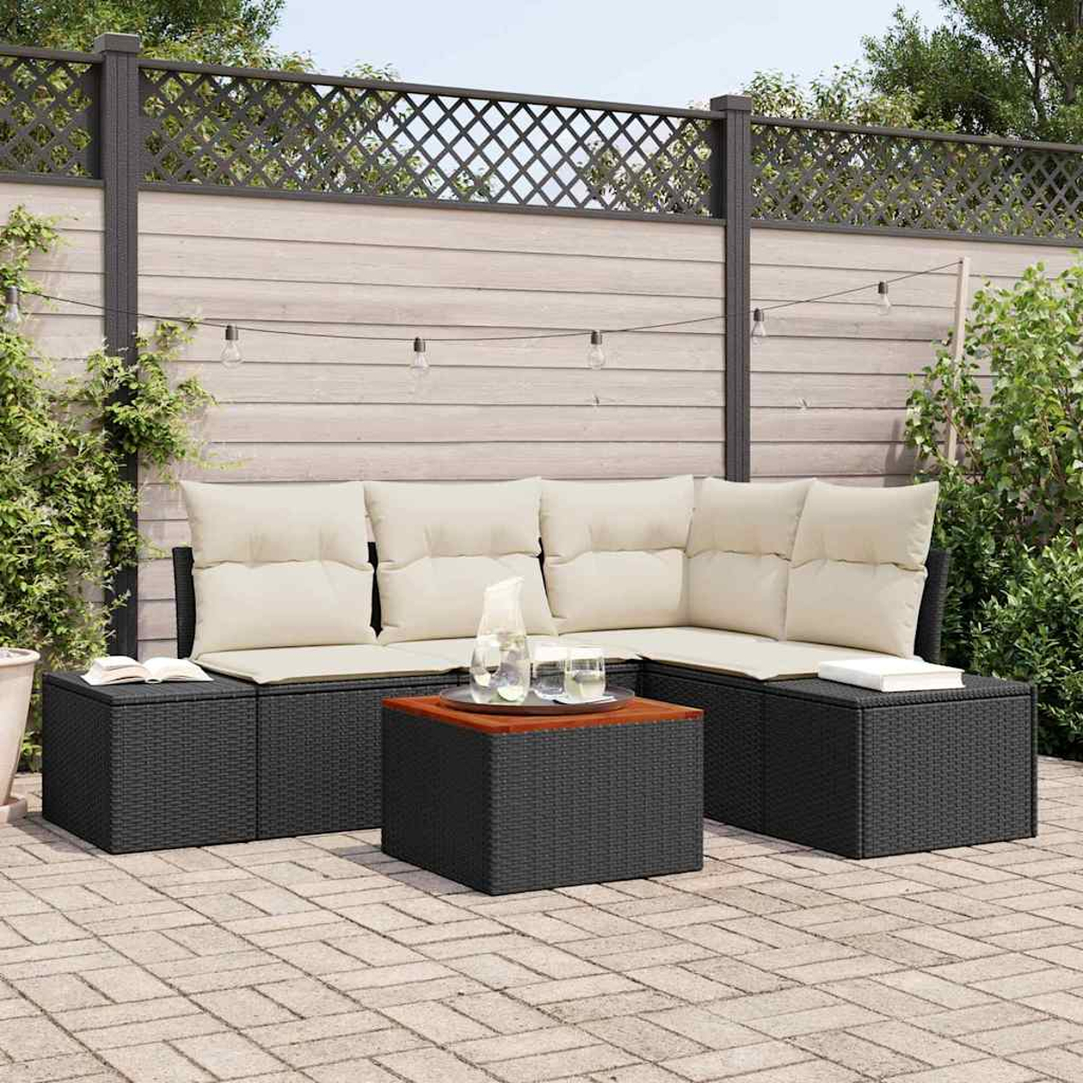 Garten-Terrassenset mit schwarzen Korbmöbeln, beigen Kissen und einem Holztisch auf einer Backstein-Terrasse. Im Hintergrund ein Holzzaun.