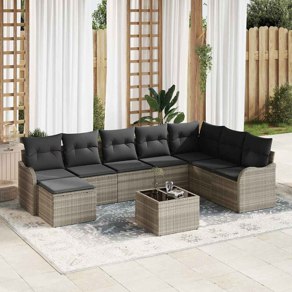 Gartensofa mit dunklen Kissen, Couchtisch und Teppich. Weiße Vorhänge und Holzzaun im Hintergrund.