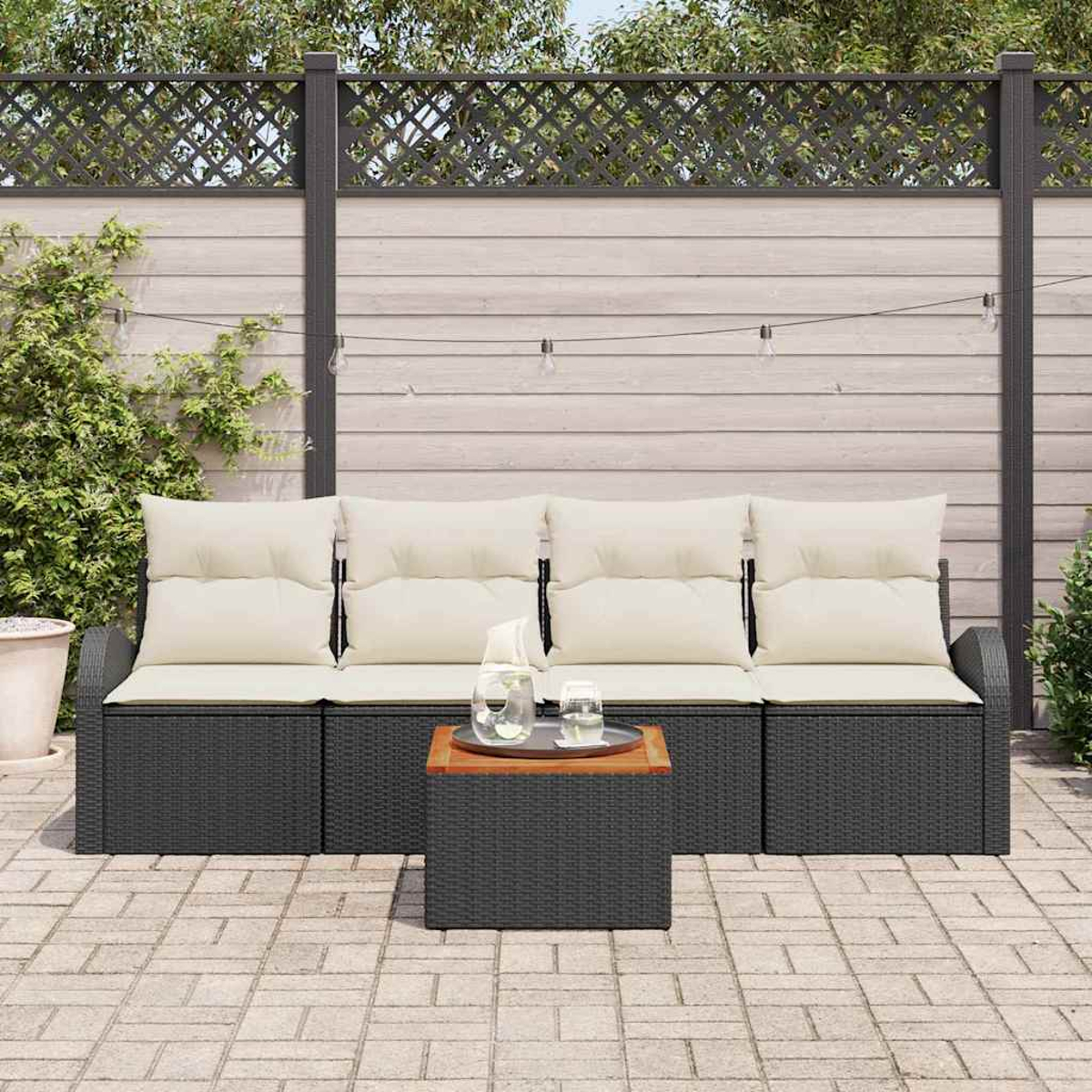 Szene im Freien mit schwarzem Rattansofa, cremefarbenen Kissen, einem Beistelltisch mit Getränken und einem Holzzaun im Hintergrund.
