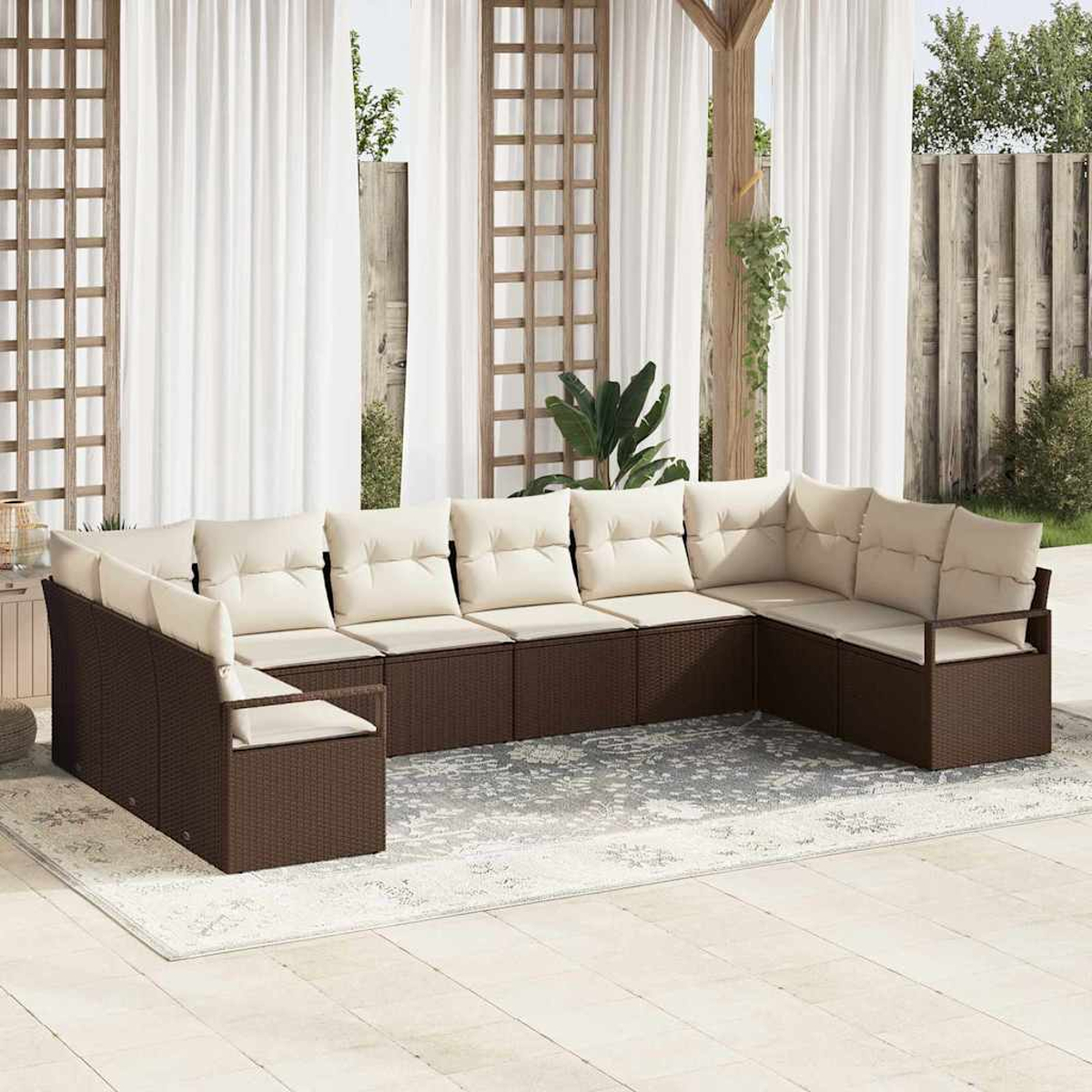 U-förmiges braunes Outdoor-Sofa aus Korbgeflecht mit cremefarbenen Kissen auf einer Terrasse mit gemustertem Teppich. Weiße Vorhänge und Holzgitter sind sichtbar.