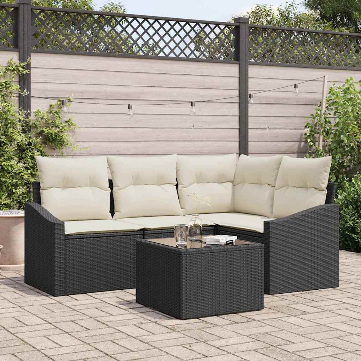 Garten-Terrassenset. Schwarzes Korbmöbel mit cremefarbenen Kissen und einem quadratischen Tisch. Dahinter ein Holzzaun und Grün.