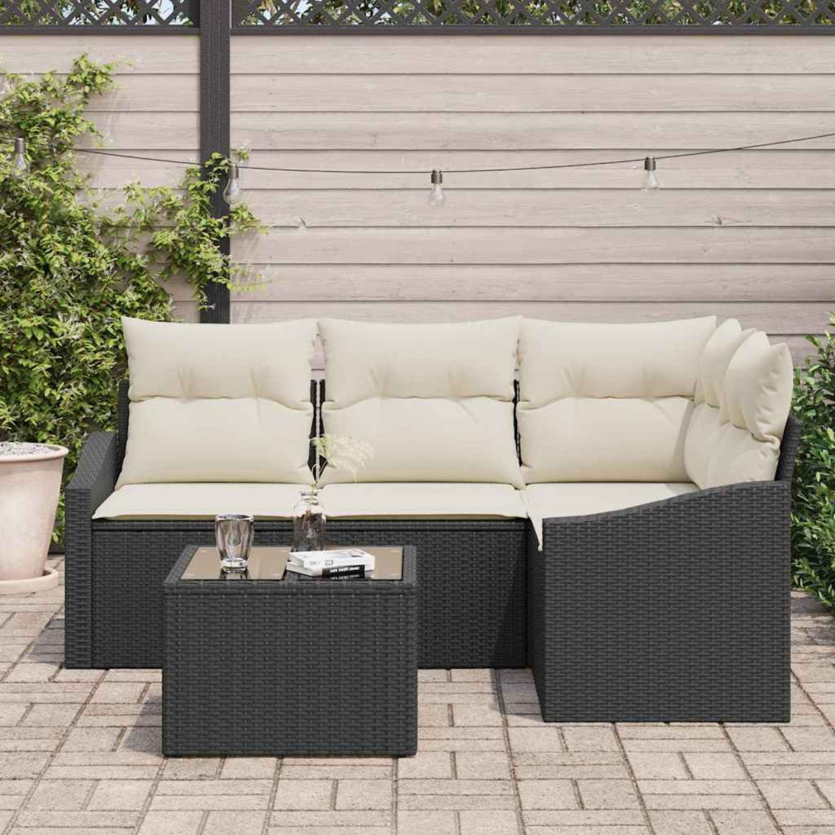 Ein dunkelgraues Outdoor-Sofa aus Korbgeflecht mit cremefarbenen Kissen und einem passenden Tisch auf einer Ziegelterrasse. Hintergrund ist ein Zaun.