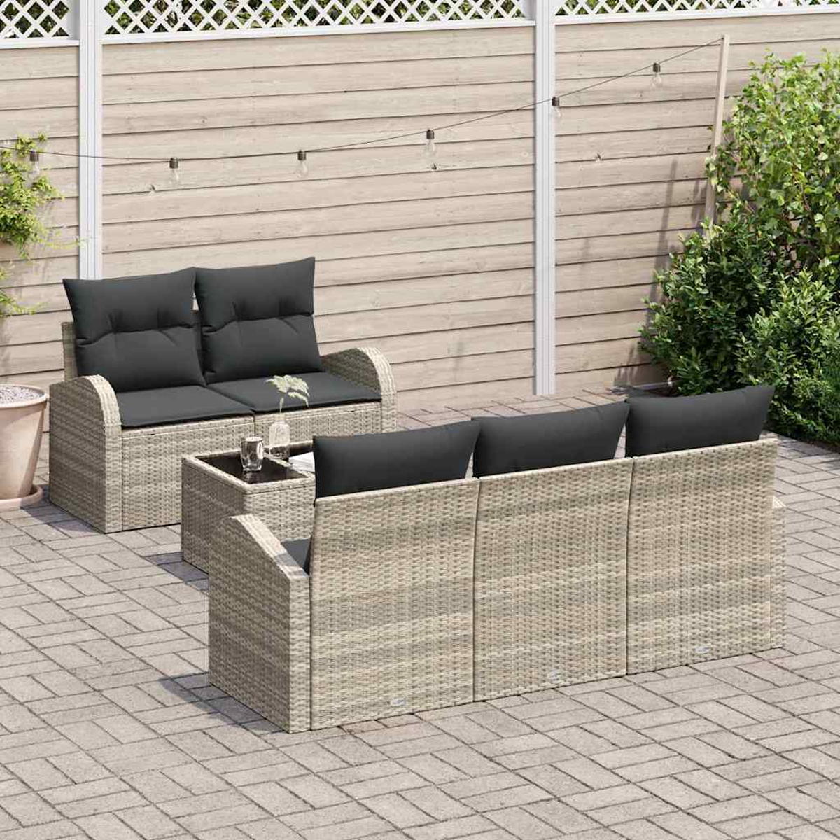 Garten-Lounge-Set: Korbsessel mit schwarzen Kissen, Glastisch und Zaun im Hintergrund. Ziegel-Terrasse mit Grün.
