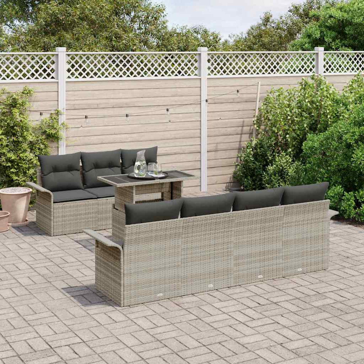 Set patio esterno con divano e divanetto marrone chiaro intrecciato con cuscini grigi, tavolino e recinzione bianca sullo sfondo.