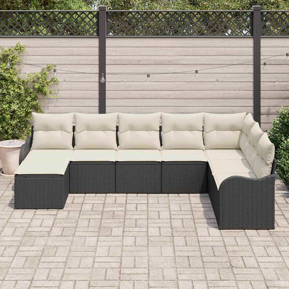 Un sofá seccional para exteriores con cojines crema y mimbre gris oscuro. Situado en un patio con valla de madera y vegetación al fondo.