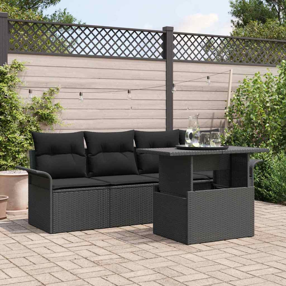 Garten-Terrassenset mit schwarzen Kissen und einem Tisch mit Gläsern und einem Krug. Holzzaun und Grün im Hintergrund.