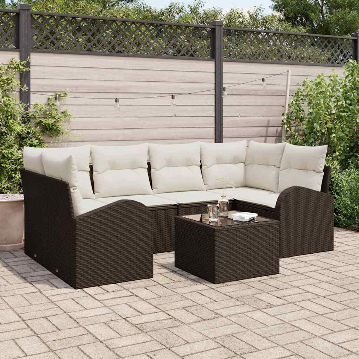 Gartenmöbel-Set aus Korbgeflecht mit cremefarbenen Kissen, einem kleinen quadratischen Tisch und Lichterketten vor einem Holzzaun.