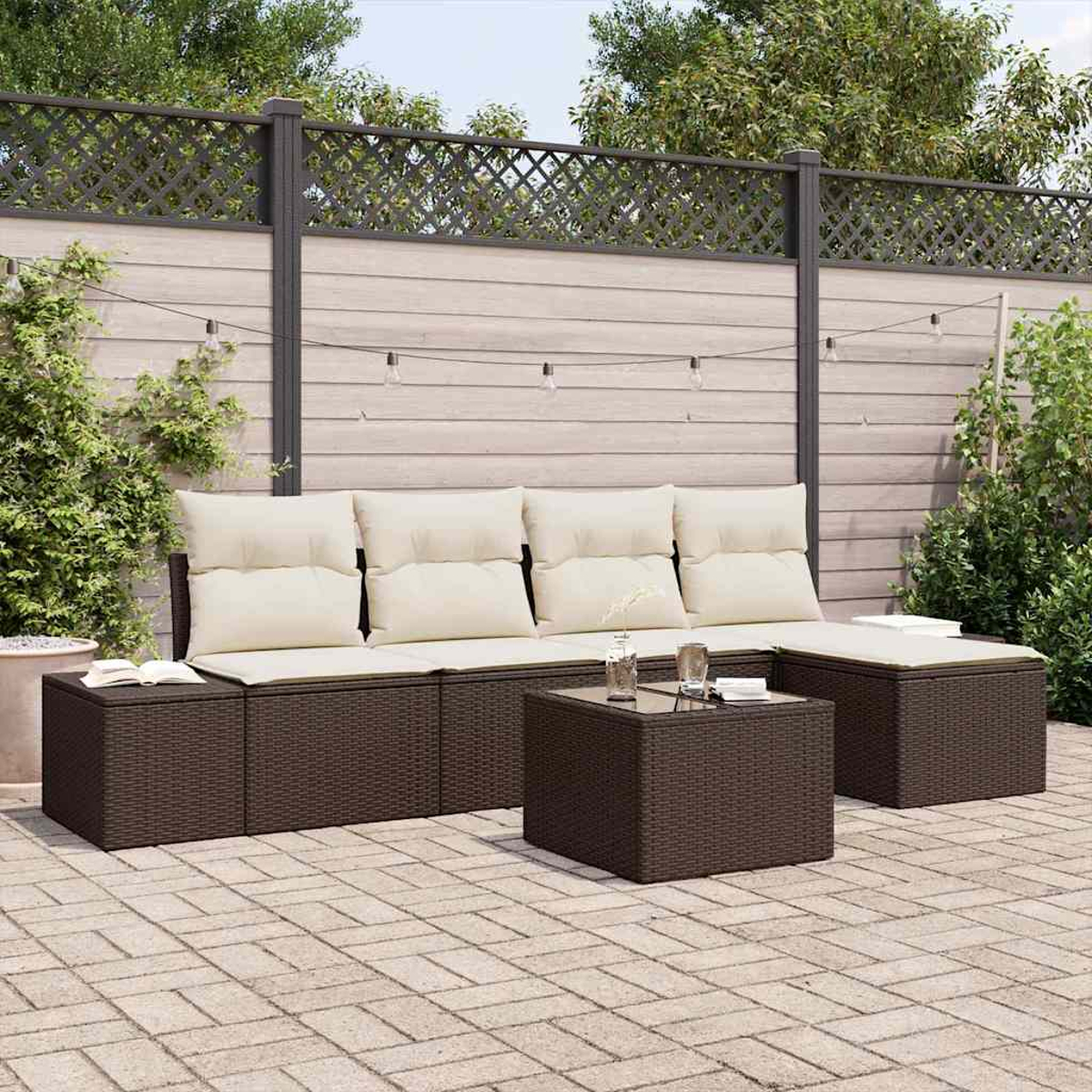 Ein braunes Outdoor-Ecksofa aus Korbgeflecht mit cremefarbenen Kissen und einem passenden Tisch auf einer Ziegelterrasse mit Zaun im Hintergrund.