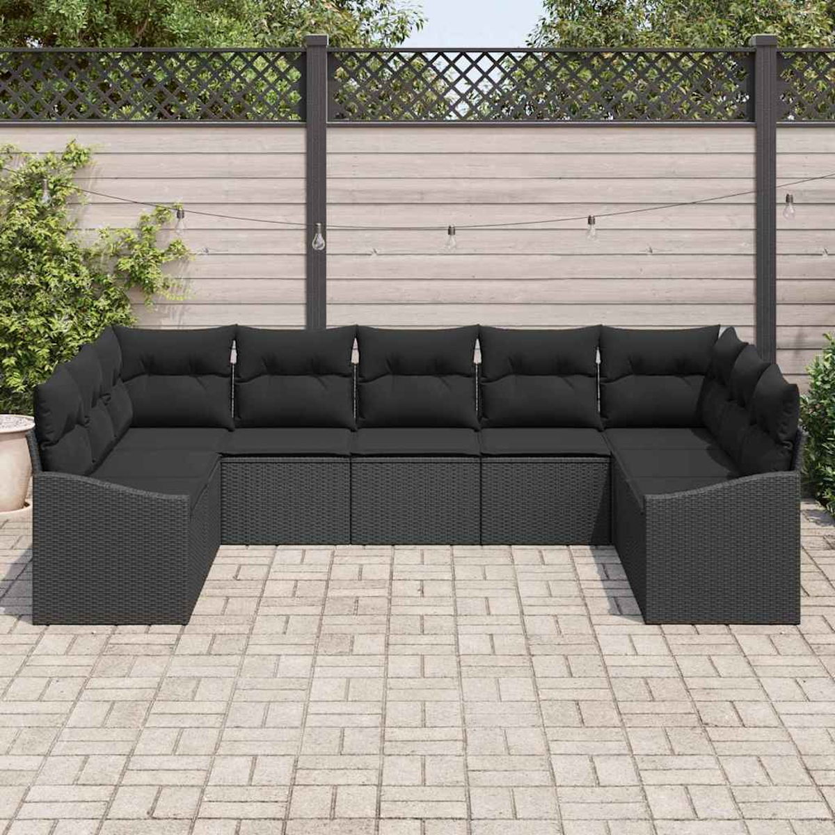 Ein U-förmiges dunkelgraues Outdoor-Sofa mit schwarzen Kissen steht auf einer gepflasterten Terrasse. Im Hintergrund ein Holzzaun und Pflanzen.