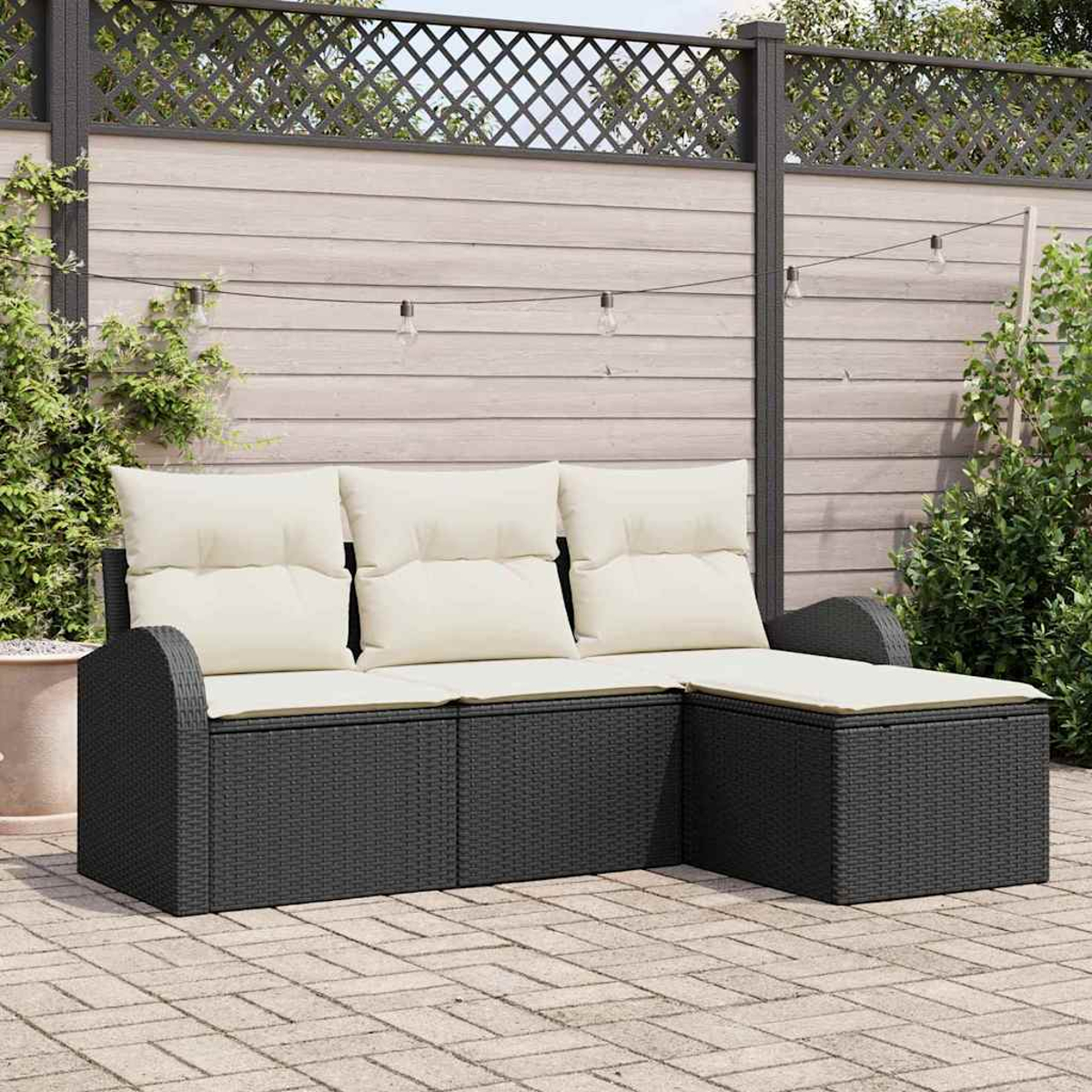 Gartencouch mit hellen Kissen. Terrasse mit Holzzaun und Lichterketten. Gepflasterte Terrasse mit Pflanzen im Hintergrund.