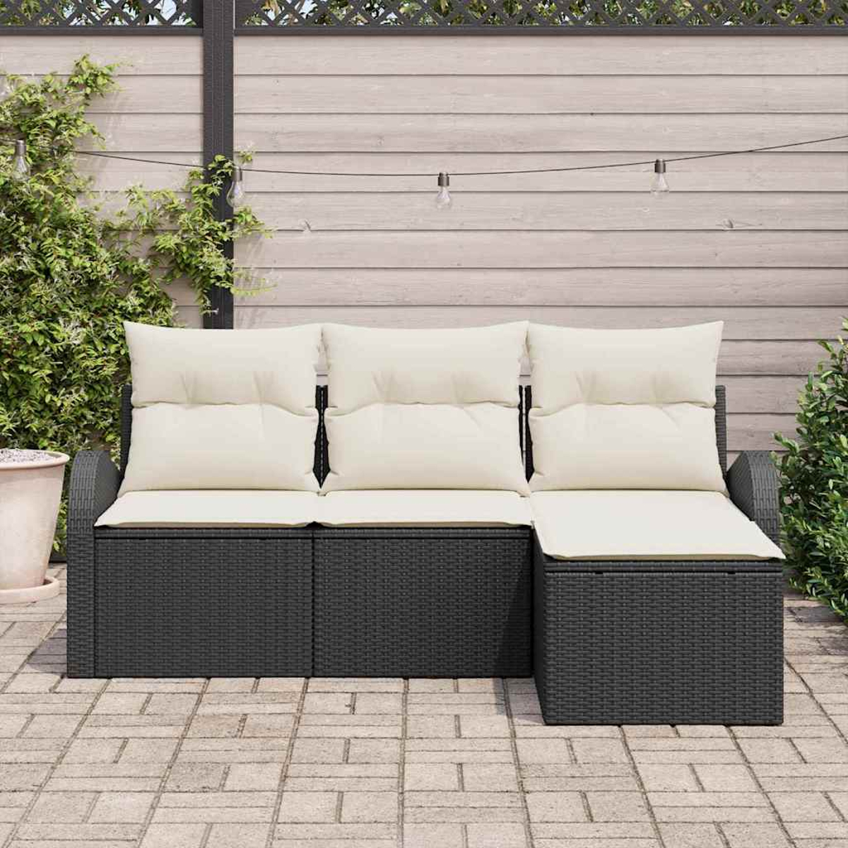 Outdoor-Ecksofa mit cremefarbenen Kissen. Dunkelgraues Korbmaterial. Beigefarbene Terrasse, heller Holzzaun und Grün im Hintergrund.