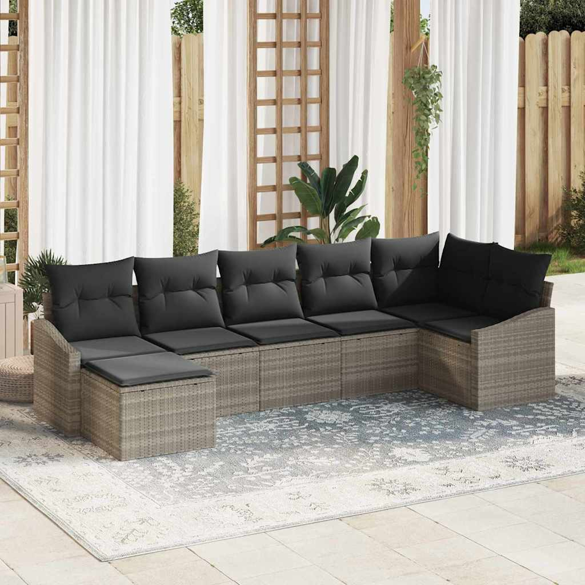 Ein Ecksofa im Freien mit grauen Kissen steht auf einem gemusterten Teppich. Weiße Vorhänge und ein Holzzaun bilden den Hintergrund.