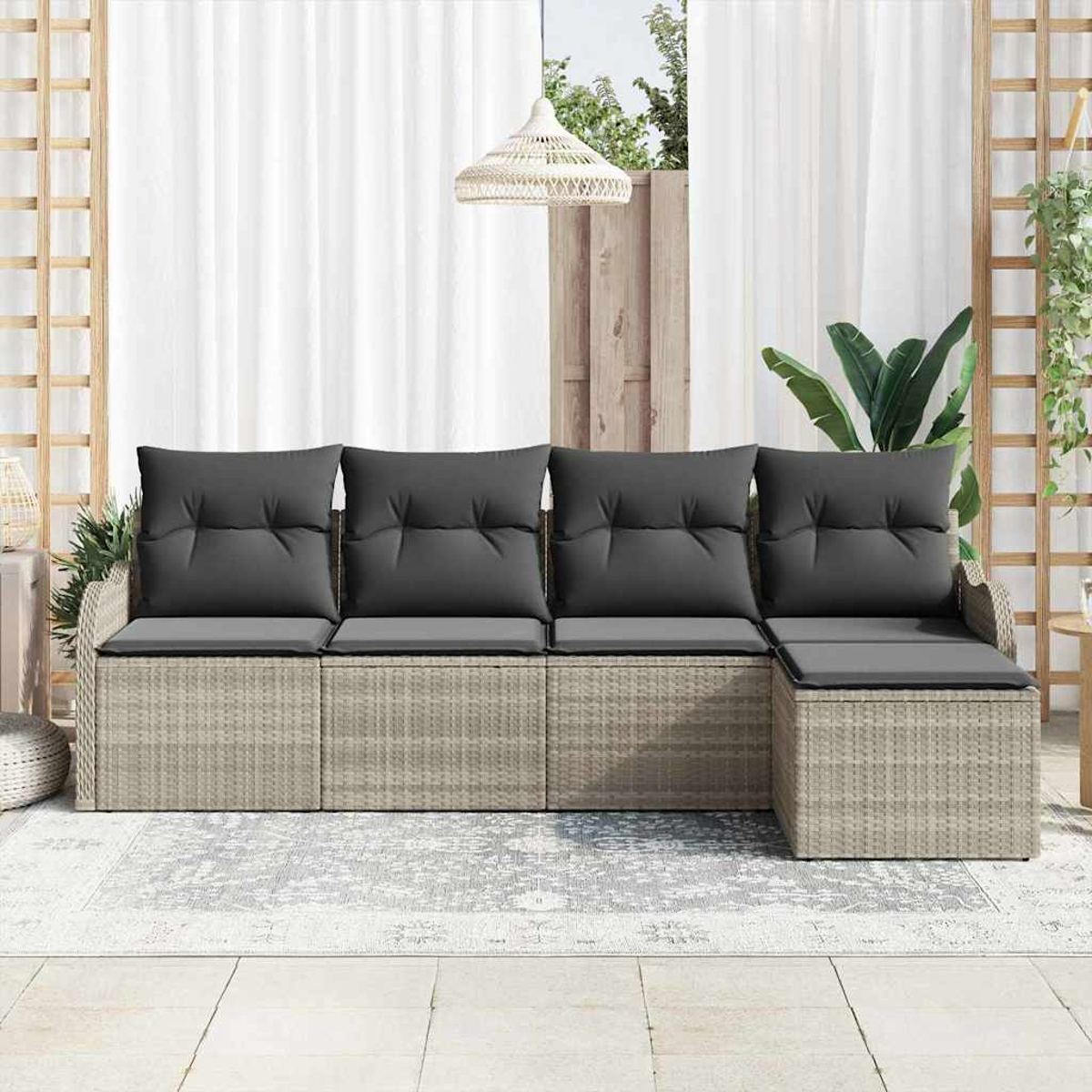 Ein graues Korbmöbelsofa mit dunkelgrauen Kissen steht auf einem gemusterten Teppich. Weiße Vorhänge und eine gewebte Lampe sind im Hintergrund.