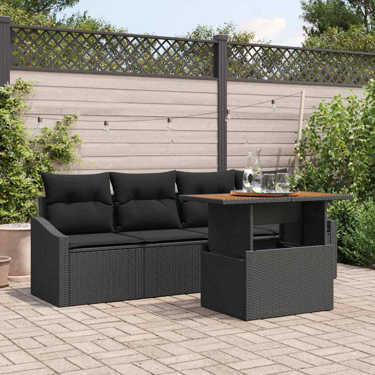 Außenszene mit einem schwarzen Korbmöbel-Sofa und Tisch. Auf dem Tisch stehen ein Krug und Gläser. Im Hintergrund ein Holzzaun.