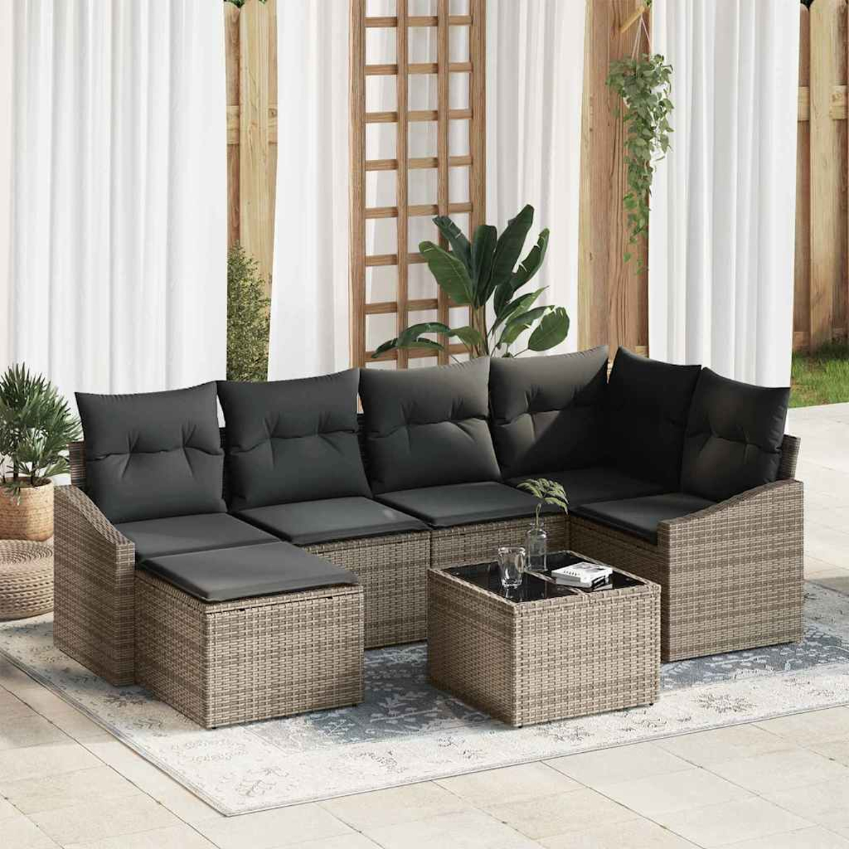 Outdoor-Korbssofa-Set mit grauen Kissen und einem kleinen Tisch auf einem Teppich. Weiße Vorhänge und Pflanzen im Hintergrund.