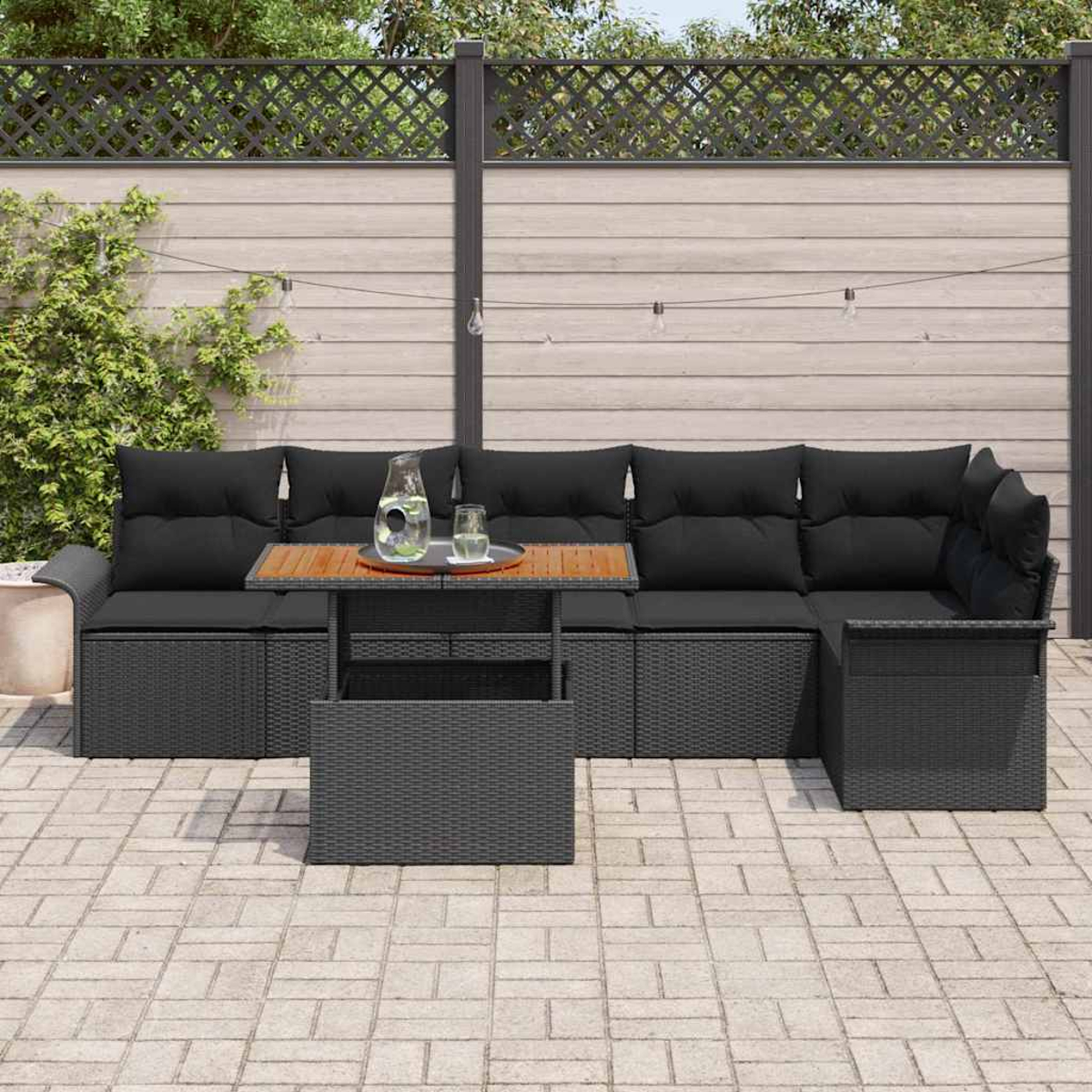 Outdoor-Ecksofa mit schwarzen Kissen und einem Holztisch, vor einem Holzzaun mit Lichterketten. Eine Kanne und ein Glas auf dem Tisch.