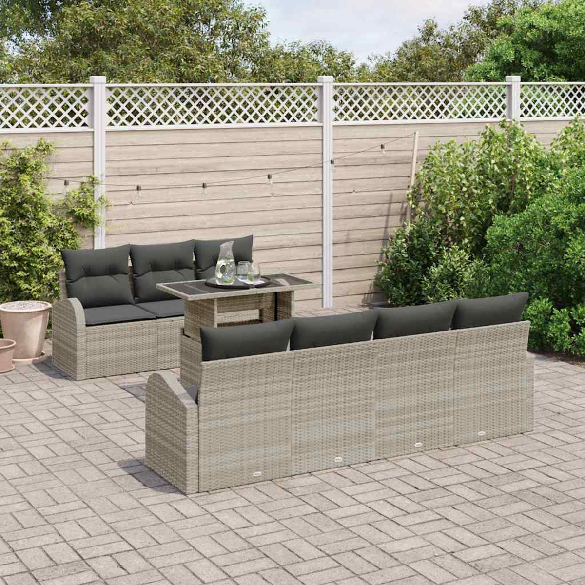 Terrassenset mit Sofa, Liebesitz und Tisch. Dunkelgraue Kissen auf grauem Korbmöbel. Weißer Zaun und Grün in der Umgebung.