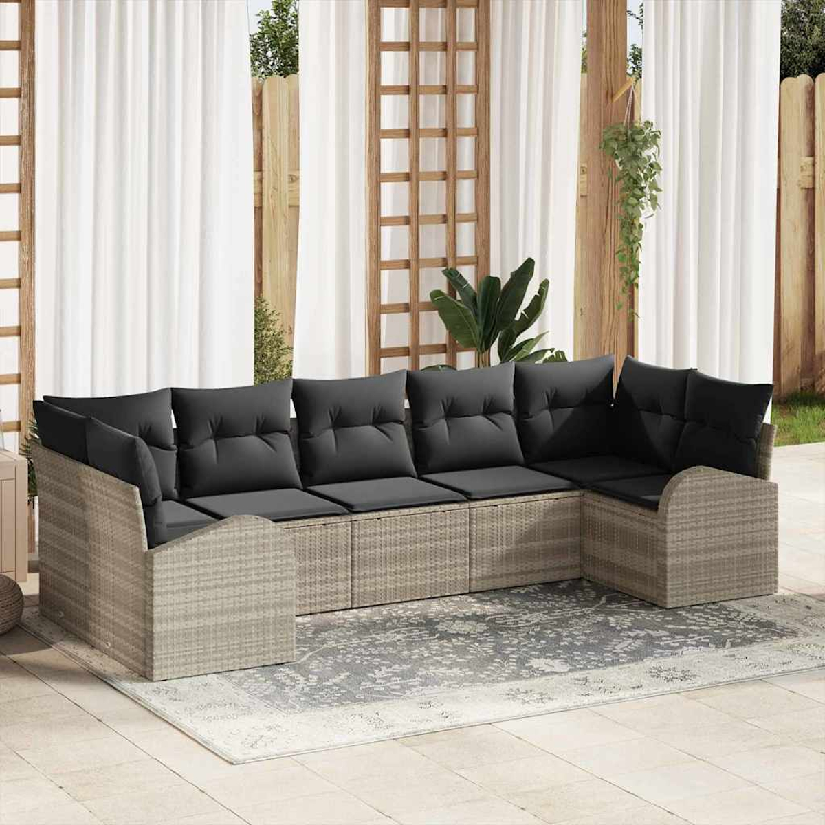 Ein geflochtenes Ecksofa mit schwarzen Kissen steht auf einem Teppich. Weiße Vorhänge und Holzbalken sind im Hintergrund.