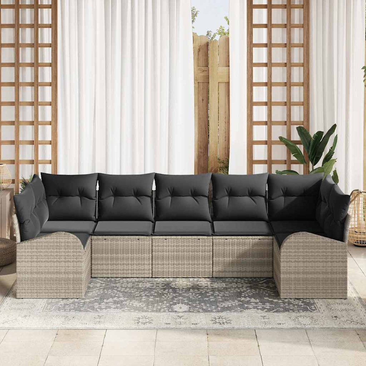 Ein graues Korbsessel-Sofa mit schwarzen Kissen steht auf einem gemusterten Teppich. Ein weißer Vorhang und ein Holzgitter sind im Hintergrund.