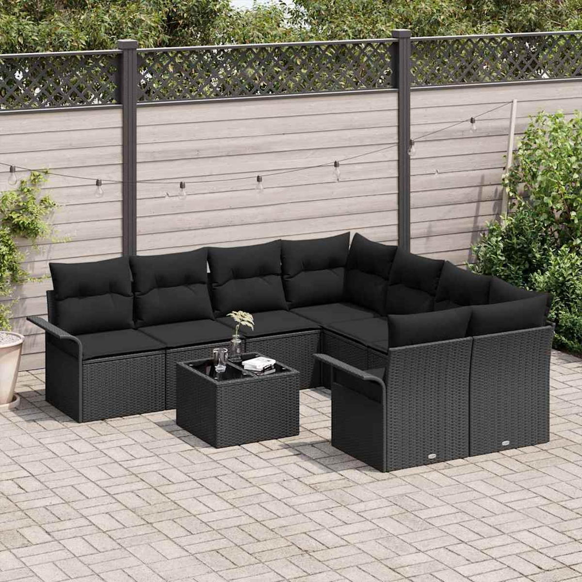 Terrassenmöbel-Set mit schwarzen Kissen und Korbmöbeln. Ein kleiner Tisch steht davor. Im Hintergrund sind ein Zaun und Pflanzen zu sehen.