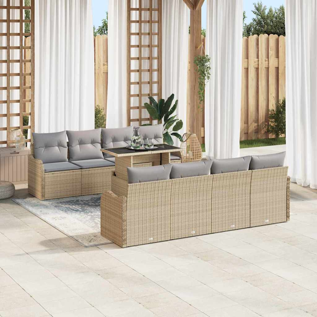 Terrassenmöbel-Set mit Korbs Sofas, grauen Kissen und einem Tisch mit Glasplatte, vor weißen Vorhängen und Holzzaun.