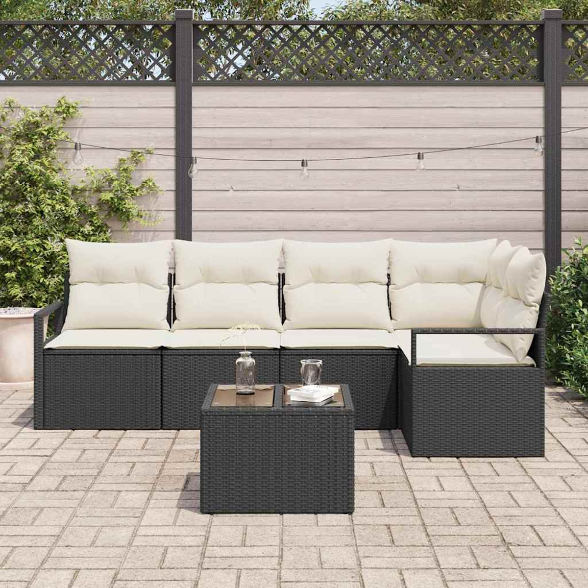 Garten-Terrassenset mit dunkelgrauen Korbmöbeln, cremefarbenen Kissen und einem Couchtisch mit Glasplatte. Hintergrund: Zaun und Grün.