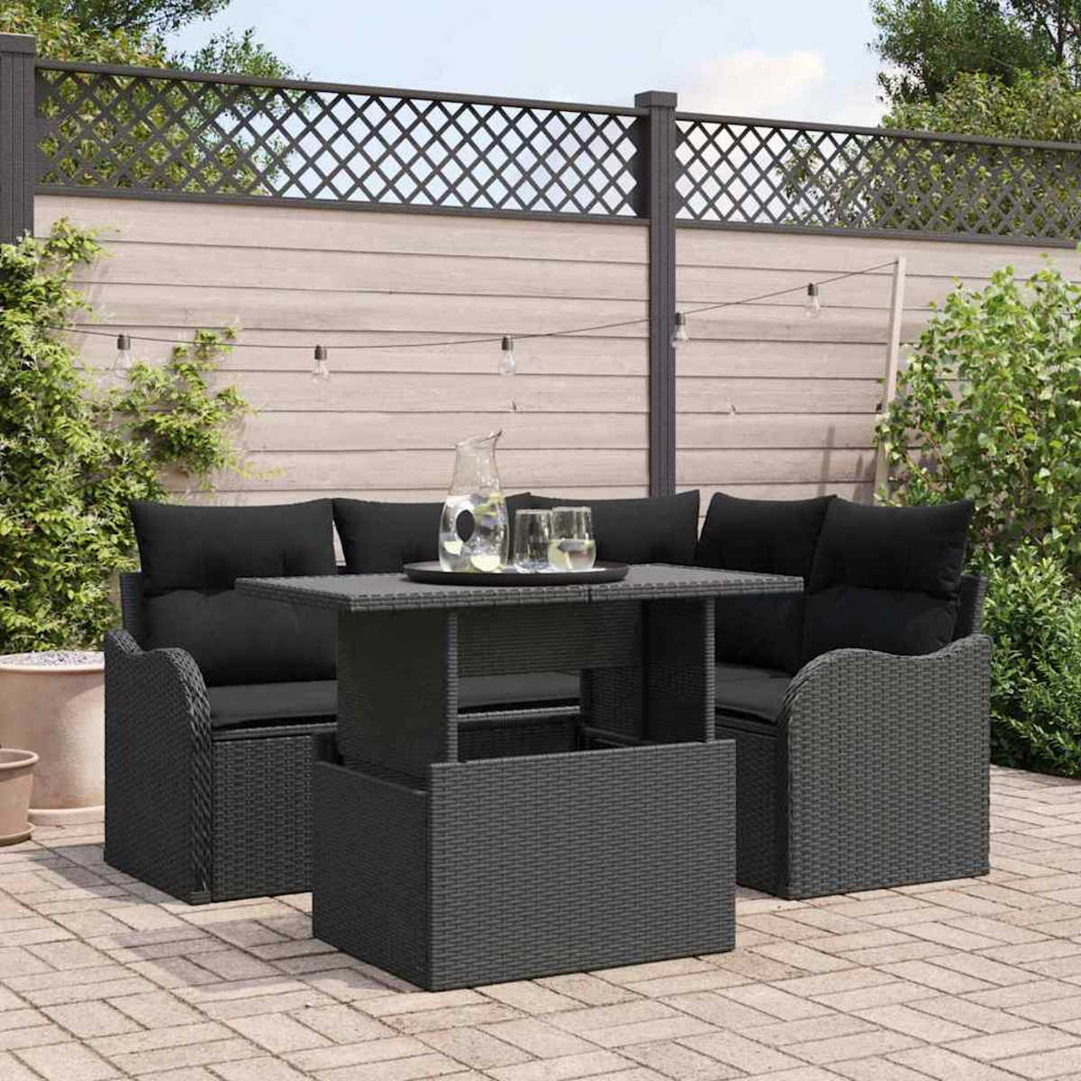 Terrassen-Set: graues Korbmöbel-Sofa und Tisch mit schwarzen Kissen. Ein Krug und Gläser stehen auf dem Tisch. Zaun und Pflanzen im Hintergrund.
