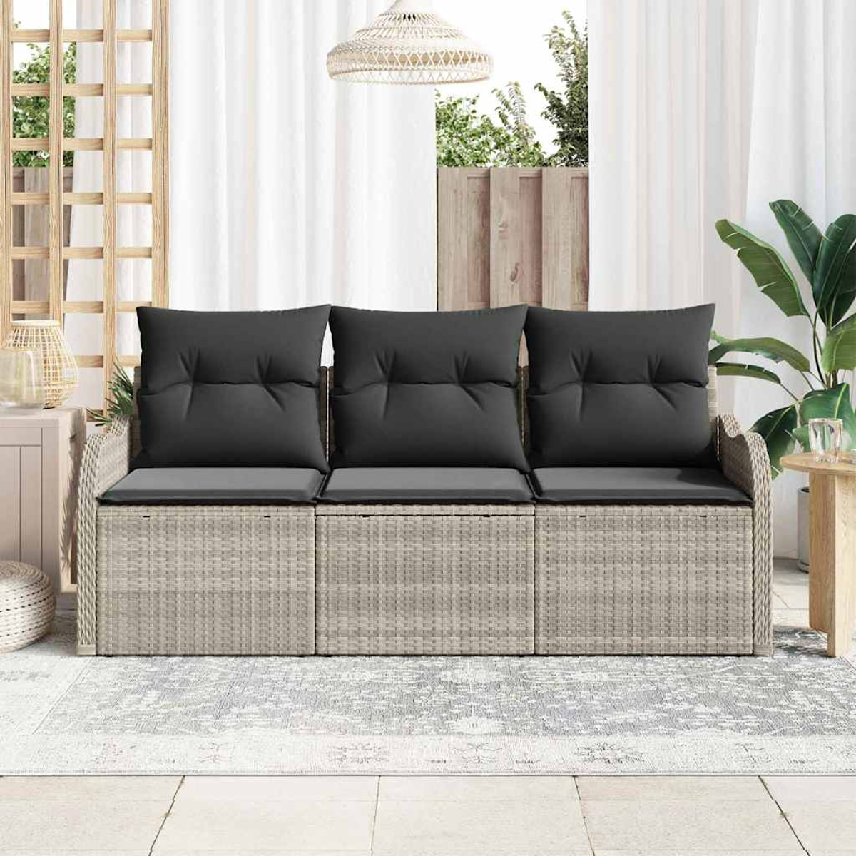 Rattansofa mit schwarzen Kissen auf der Terrasse. Weiße Vorhänge, gewebte Lampe und grüne Pflanzen im Hintergrund.