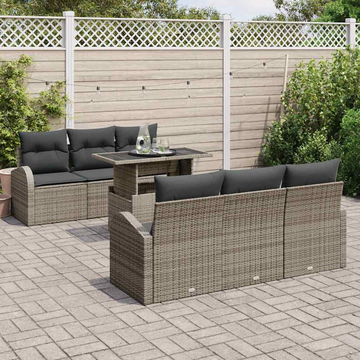 Terrassen-Set im Freien. Graue Korbsessel und -tisch. Schwarze Kissen und ein Glastisch. Hintergrund: Holzzaun und grüne Büsche.