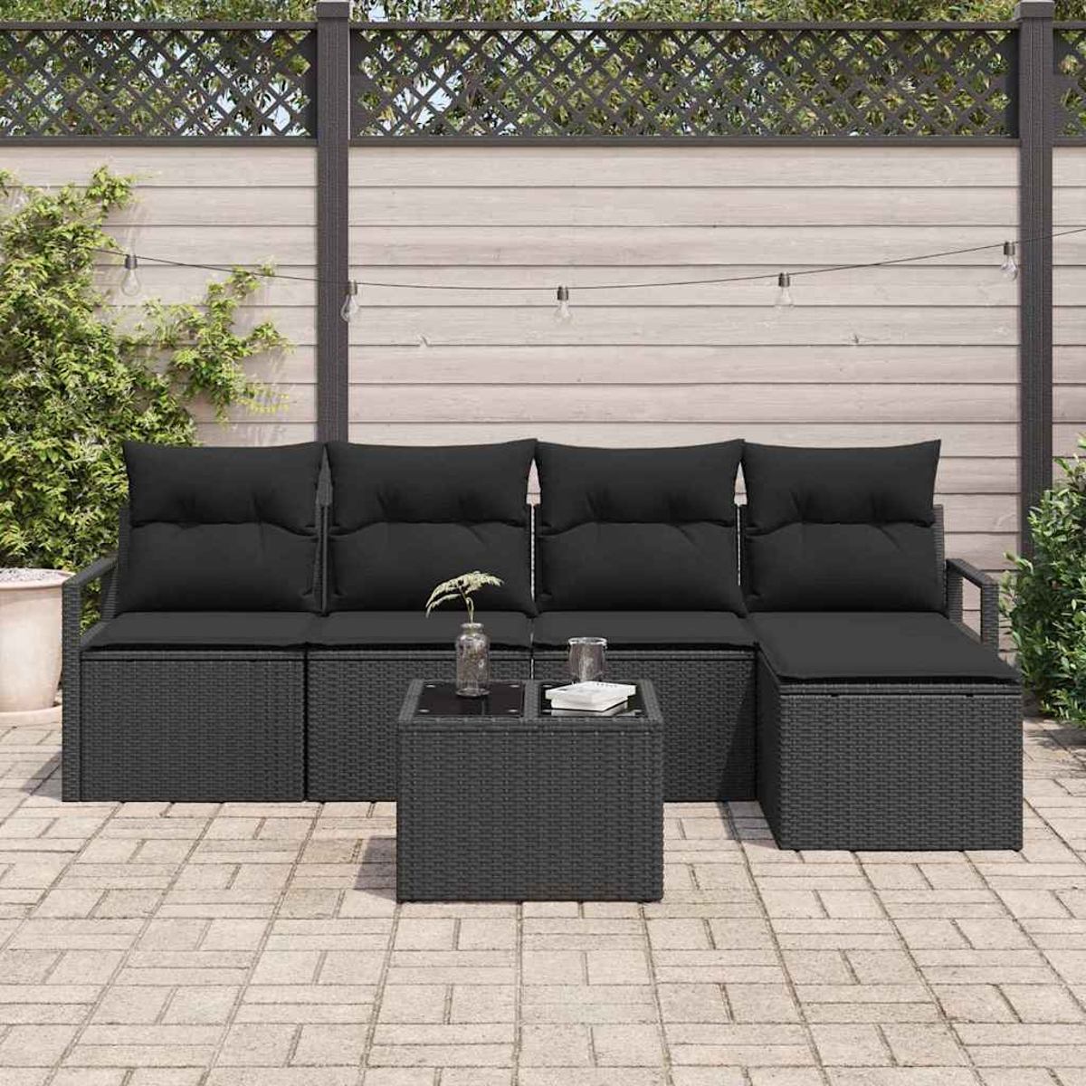 Schwarzes Outdoor-Rattan-Sofaset mit Kissen, Couchtisch und Lichterketten auf einer gepflasterten Terrasse mit Zaun.