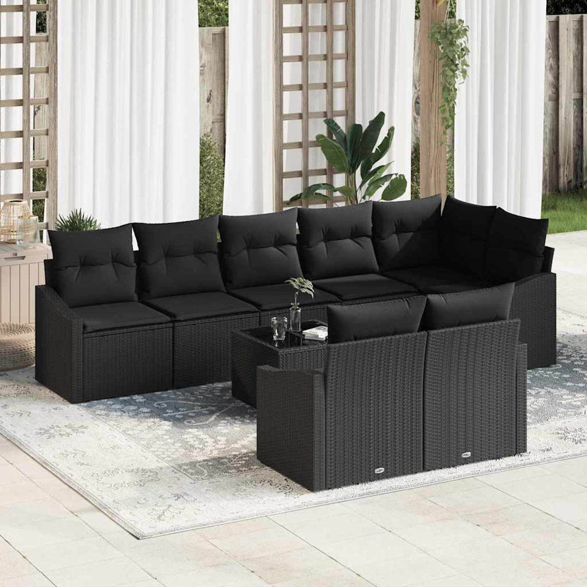 Schwarzes Outdoor-Geflecht-Ecksofa mit Kissen, kleinem Tisch und Teppich auf einer Terrasse. Weiße Vorhänge und Grün im Hintergrund.